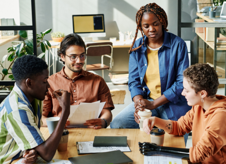 Four young adults gather around a clipboard and some papers. One has dark skin and short, curly hair. One has medium-light skin and dark hair. One has dark skin and long braids. One has light skin, short brown hair and a prosthetic arm.
