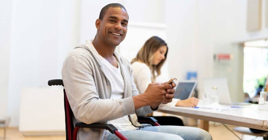 Photo: Person with dark skin in a wheelchair wearing casual clothing is smiling while working in an office setting.