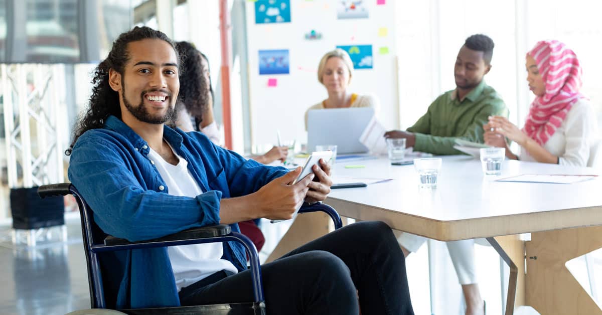 A man in a blue shirt using a wheelchair is at a table with colleagues in an office.