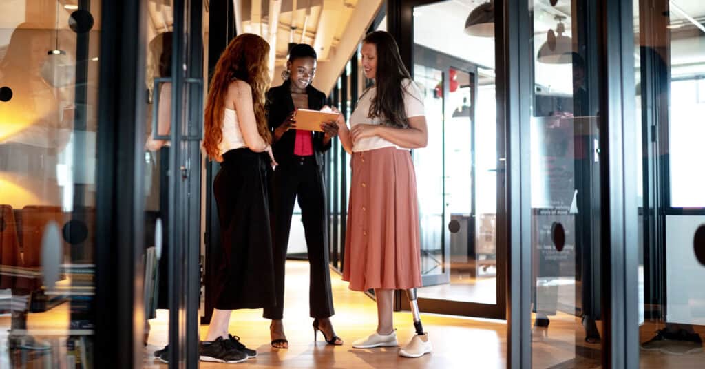 Trois femmes sourient et regardent une tablette dans un bureau.