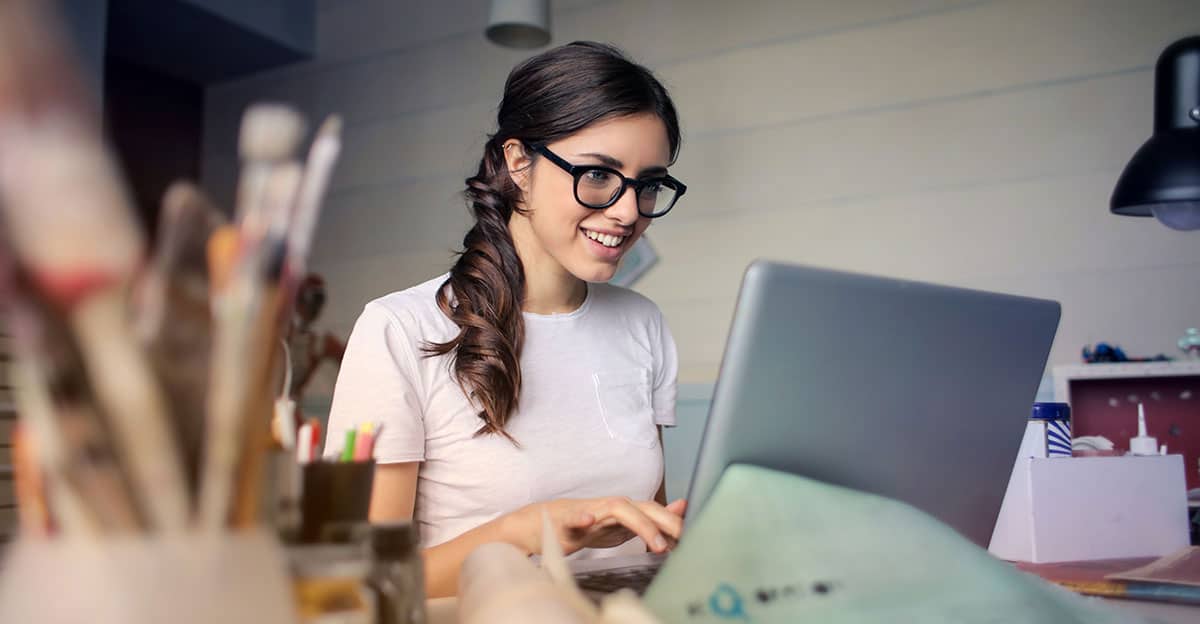 Person with long hair and glasses sitting at a desk and looking at a laptop.