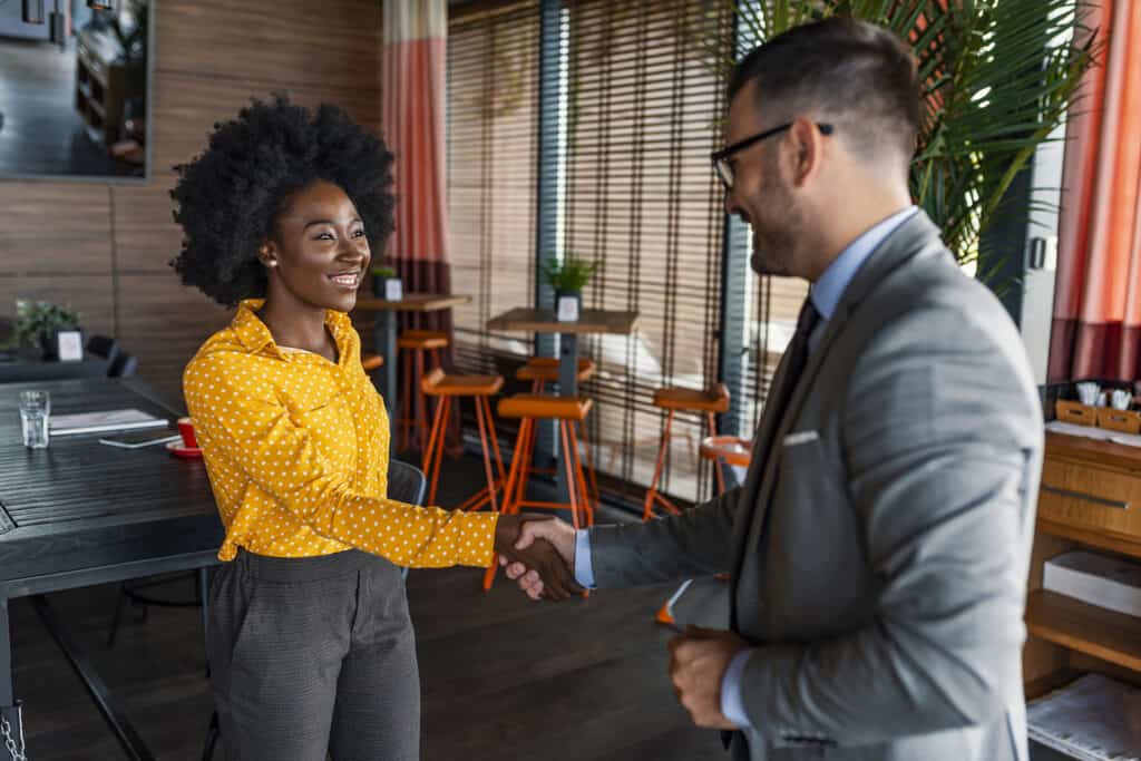 Deux personnes portant des vêtements de travail se tiennent debout et se serrent la main, avec de petites tables basses et des chaises en arrière-plan. Cette image représente un développeur d'emplois en contact avec un employeur.