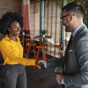 Two people wearing business clothing are standing and shaking hands, with small coffee tables and chairs in the background. This image represents a job developer connecting with an employer.