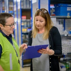 In a warehouse, an employer and employment practitioner review a document on a clipboard. This image represents a job-site analysis (JSA), one of the tools discussed in the Supported Employment Essentials program.