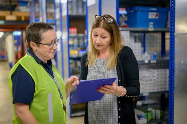 Dans un entrepôt, un employeur et un professionnel de l'emploi examinent un document sur un presse-papiers. Cette image représente une analyse de chantier (JSA), l'un des outils abordés dans le programme Supported Employment Essentials.