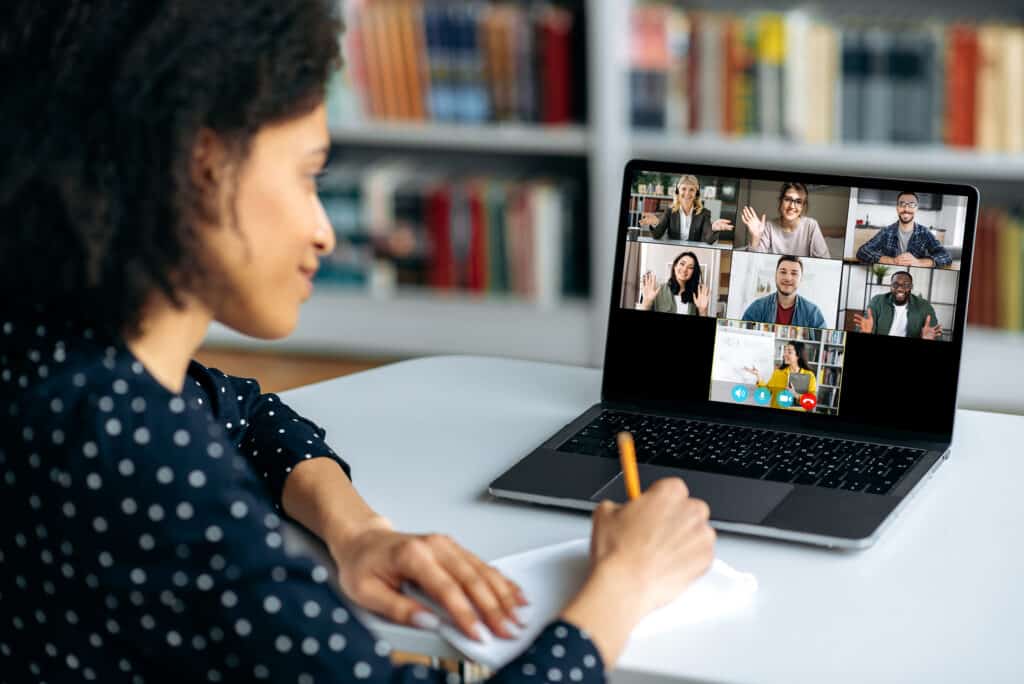 A person is smiling while writing notes and attending a virtual group meeting on a laptop.