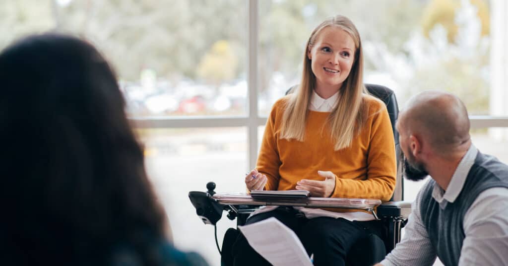 Photo : Personne en tenue professionnelle en fauteuil roulant avec une tablette parlant à d'autres personnes sur un lieu de travail