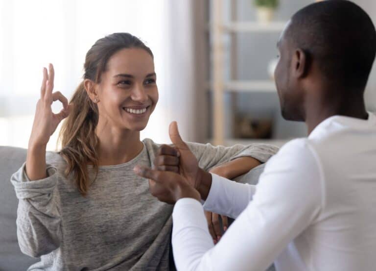 Photo: Two people, one with medium-dark skin and long brown hair and one with dark skin and very short dark hair, are using sign language.