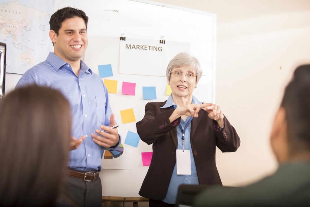 An ASL interpreter stands beside the meeting facilitator at a work meeting. There are sticky notes on a whiteboard behind them.