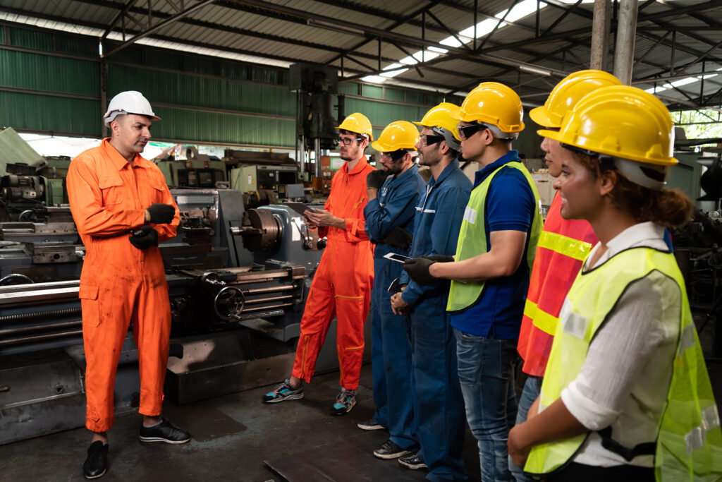 In a factory, a group of employees wearing safety gear attend a meeting with their supervisor.