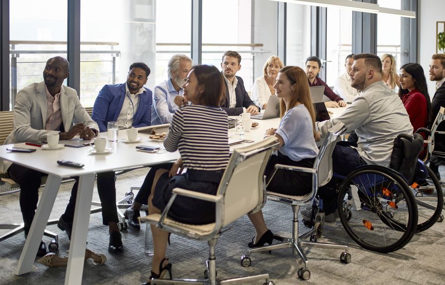 Diverse employees, including a person in a wheelchair, sit around a table in an office.