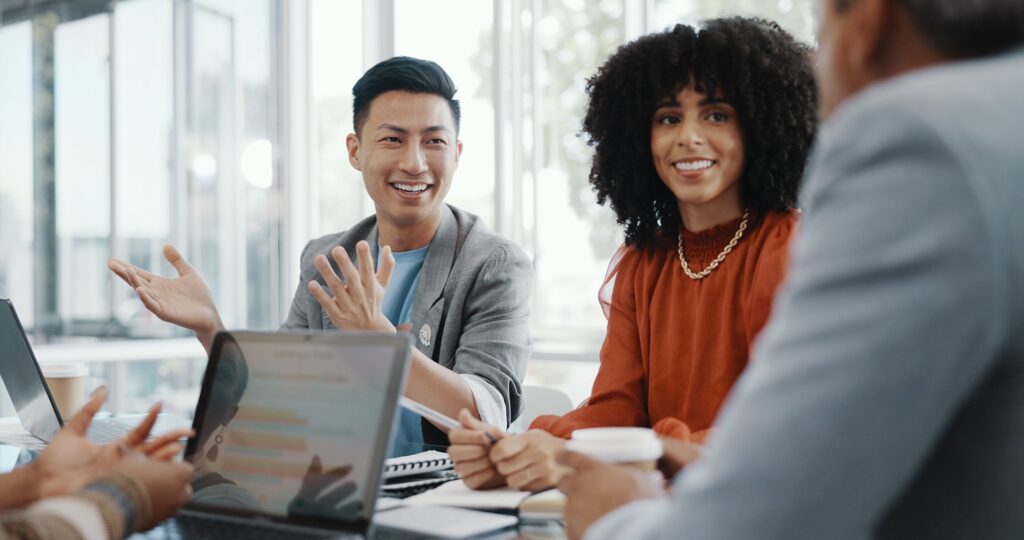 In a meeting room, a group of people with different racial identities sit and have a discussion. The image depicts the importance of meaningful collaboration between different individuals to boost workplace diversity, equity, and inclusion.