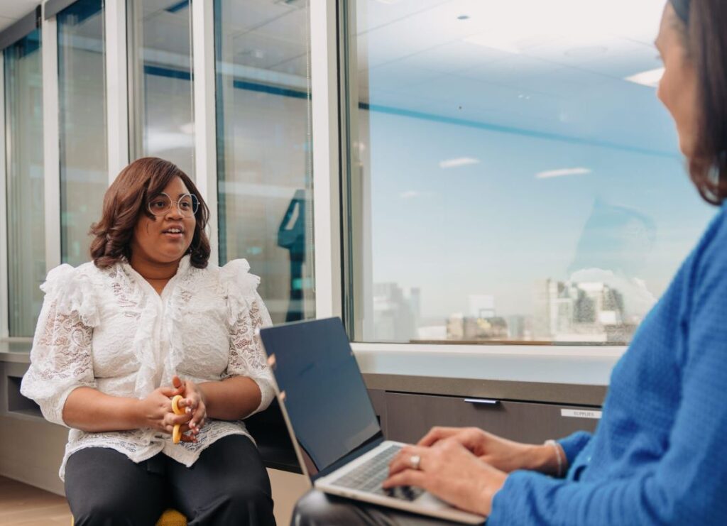 A medium-skinned young woman holding a fidget toy sits in an office during a meeting with her mentor who is medium-skinned and who is typing on a laptop.