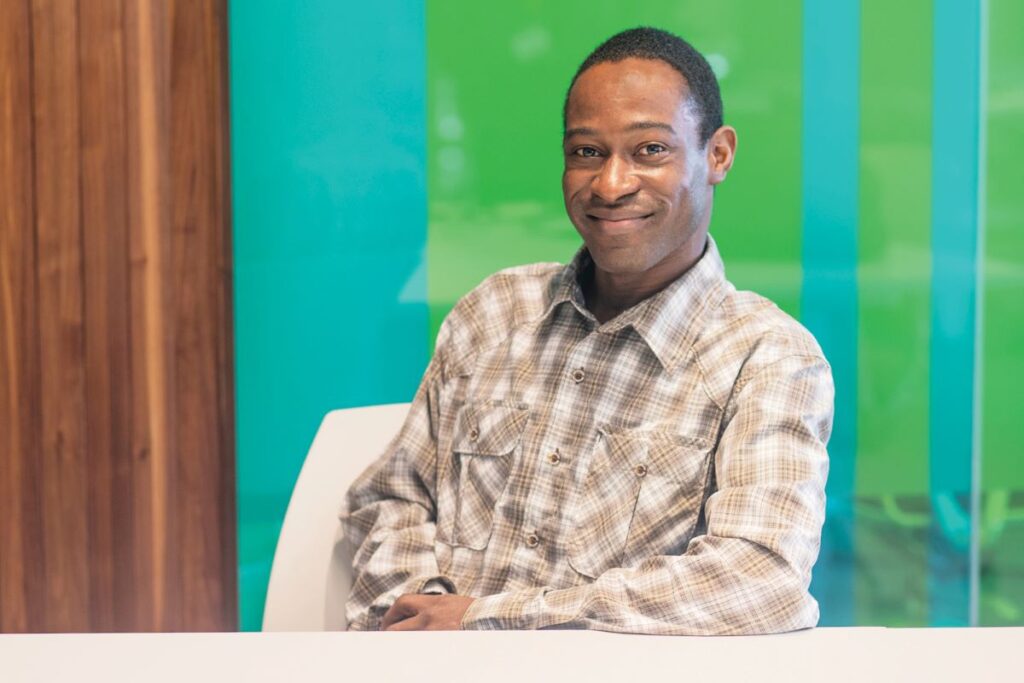 Photo: Dr. De-Lawrence Lamptey. Person with dark skin and short dark hair and brown eyes smiling, wearing white and light brown plaid shirt, sitting with teal and bright green vertical lines on wall behind him.