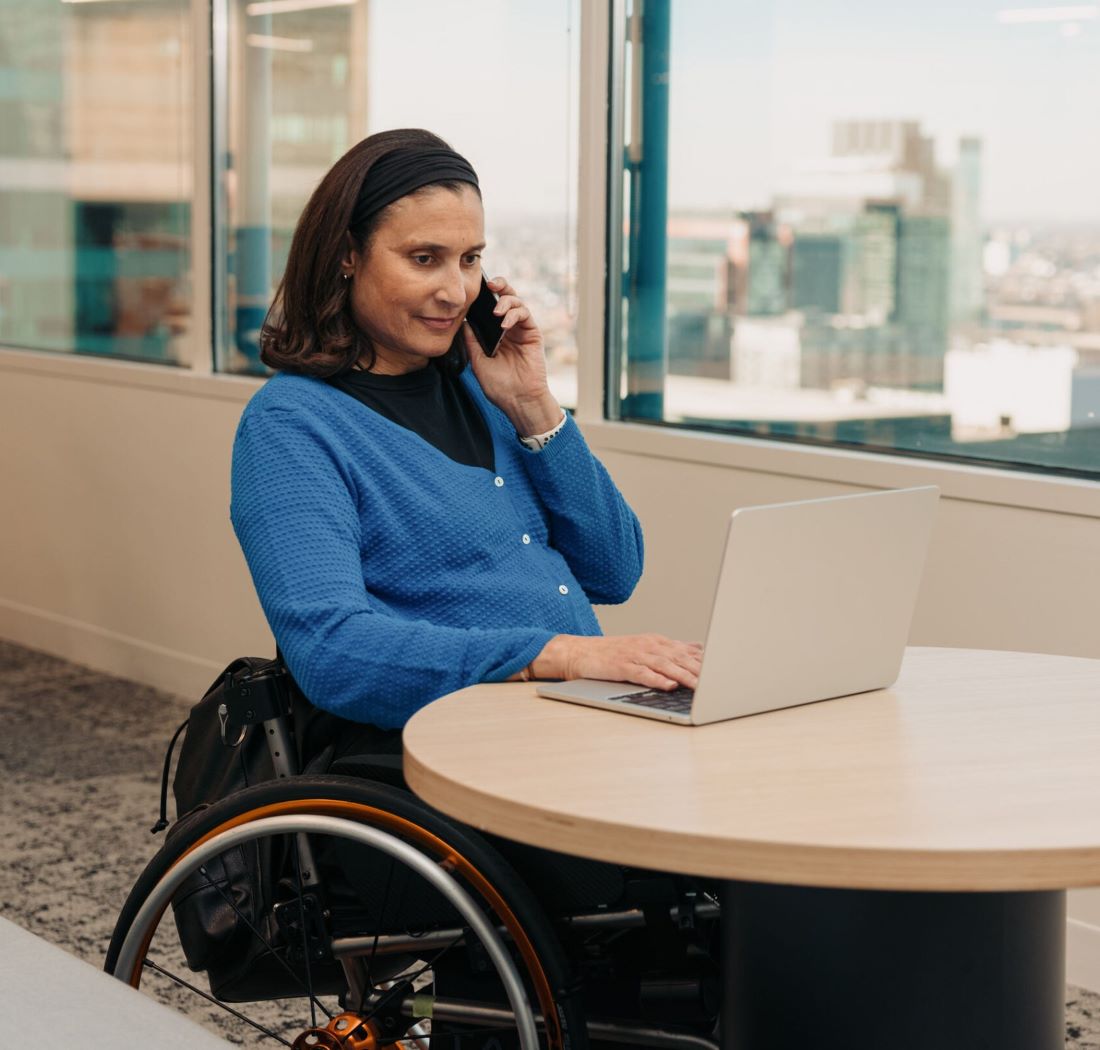 Woman with medium skin tone and shoulder length dark hair is wearing a blue sweater. She is sitting in a wheelchair at a round table in an office setting, looking at a laptop and speaking on a cellphone.