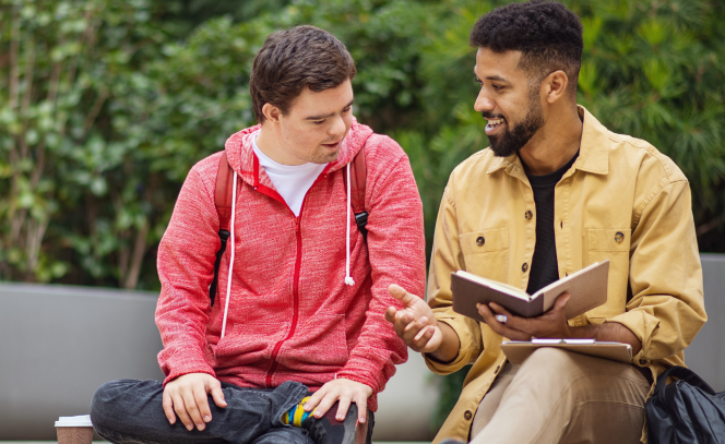Two people discuss an open book. Both have short, dark hair. One has light skin and wears a red sweatshirt. One has medium-dark skin and a beard and wears an unbuttoned yellow shirt over a black t-shirt.