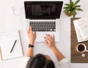 A person with medium-light skin tone uses a laptop with papers lying on the table around them.