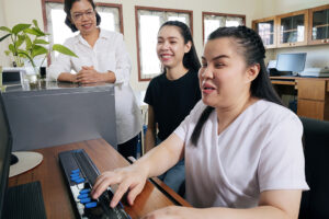 Vue grand angle de collègues asiatiques heureuses dans un bureau, y compris une personne atteinte de cécité utilisant un ordinateur doté d'un dispositif d'assistance à affichage braille actualisable. Inclusion du handicap.