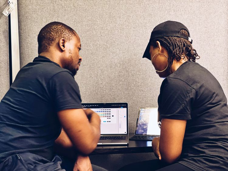 Two people with medium skin tone, a mentor and a mentee, are sitting at a desk and looking at a laptop.