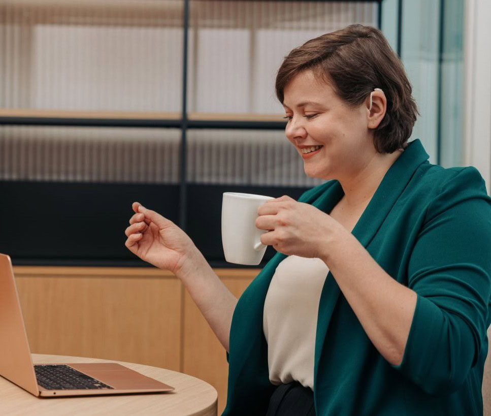 A professional works on their laptop while holding a cup of coffee in an office. They have their hair tucked behind their ear to show off their hearing aid.