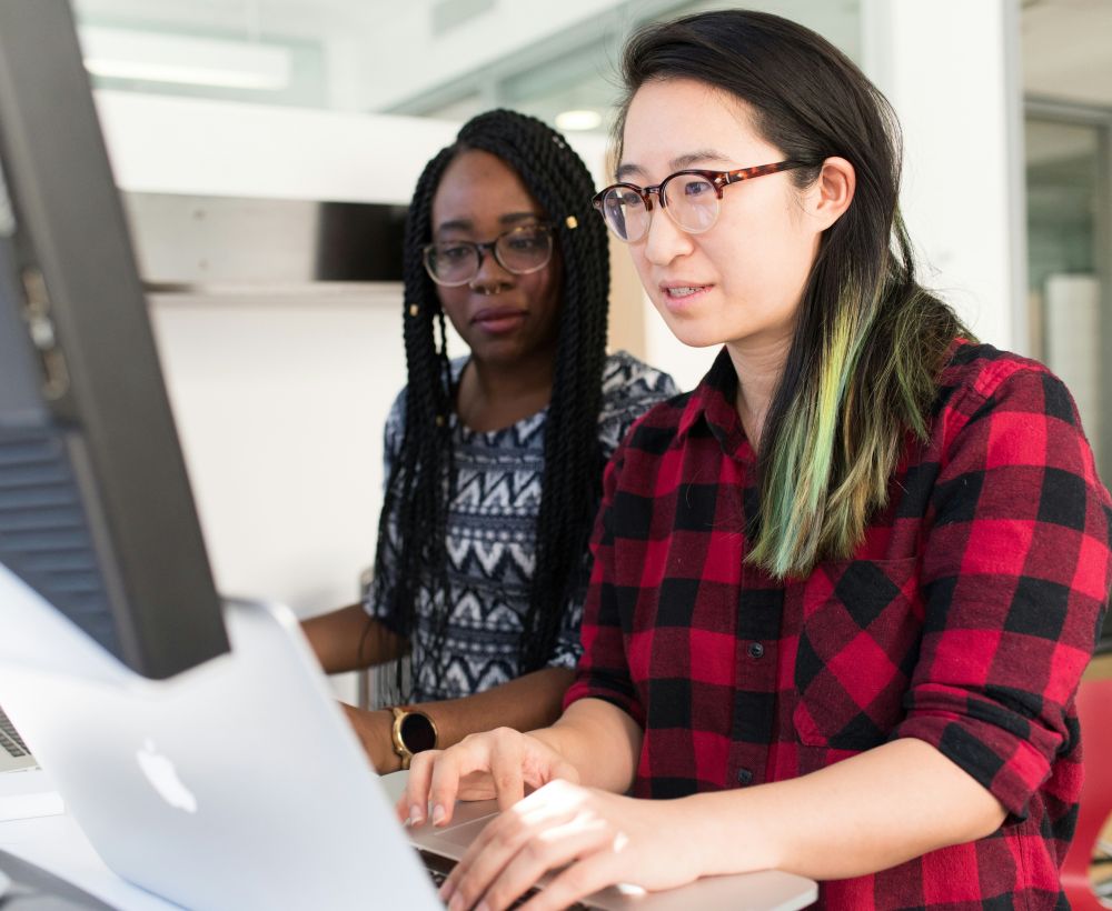 Two racialized people are sitting at a desk, one is typing on a laptop and looking at a larger monitor and the other is looking at the laptop.