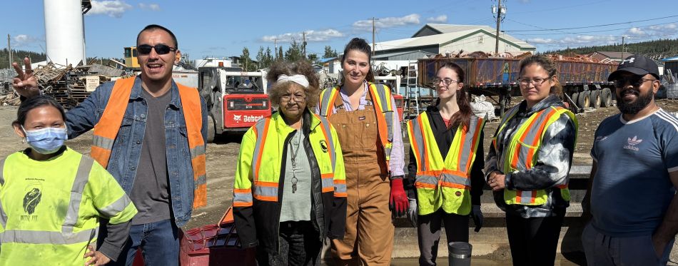 A group of people wearing safety vests smile as they stand in a recycling centre.
