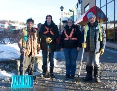 Four people stand smiling outside on a city sidewalk in the winter holding shovels and other tools.