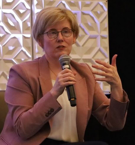 Photo of a person with short blond hair and glasses wearing a salmon pink blazer and cream blouse, who is speaking into a mic.