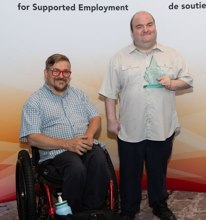 Photo of Phillip Emmerson, who uses a wheelchair, and Scott MacLellan, who is holding an award made of clear glass with a green tinge.