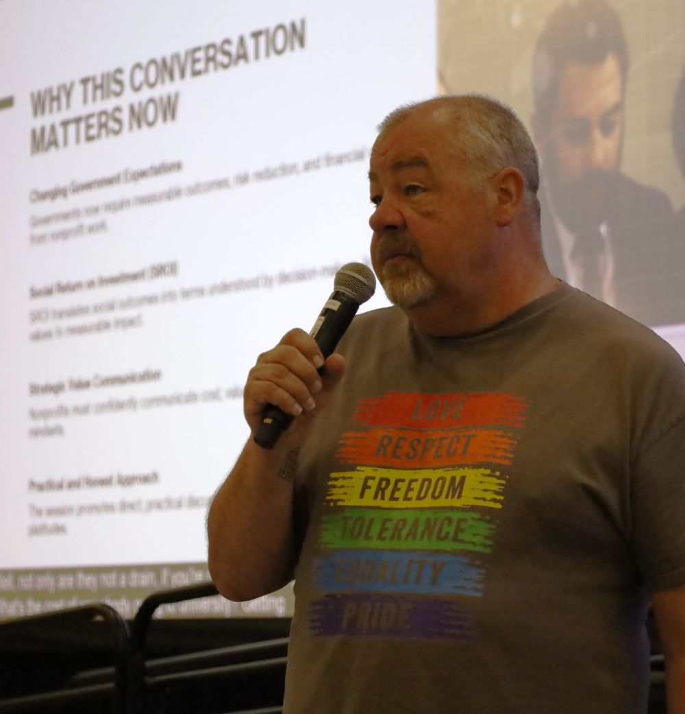 Photo of a person with short salt and pepper hair, holding a mic and wearing a t-shirt with “Love – Respect – Freedom – Tolerance – Equality – Pride.” A large screen in the back says, “Why this conversation matters now.”