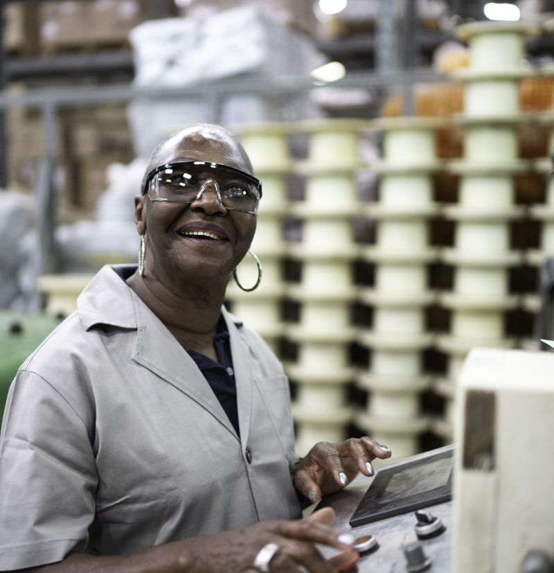 A person smiles at the camera as they work in a factory. They wear protective glasses as they stand at a terminal pushing buttons.
