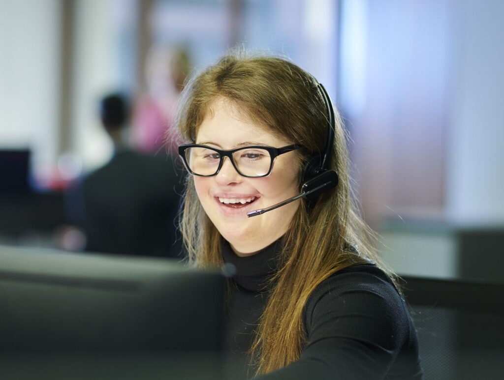 A person with Down syndrome wears a microphone and is working on a computer in an office.