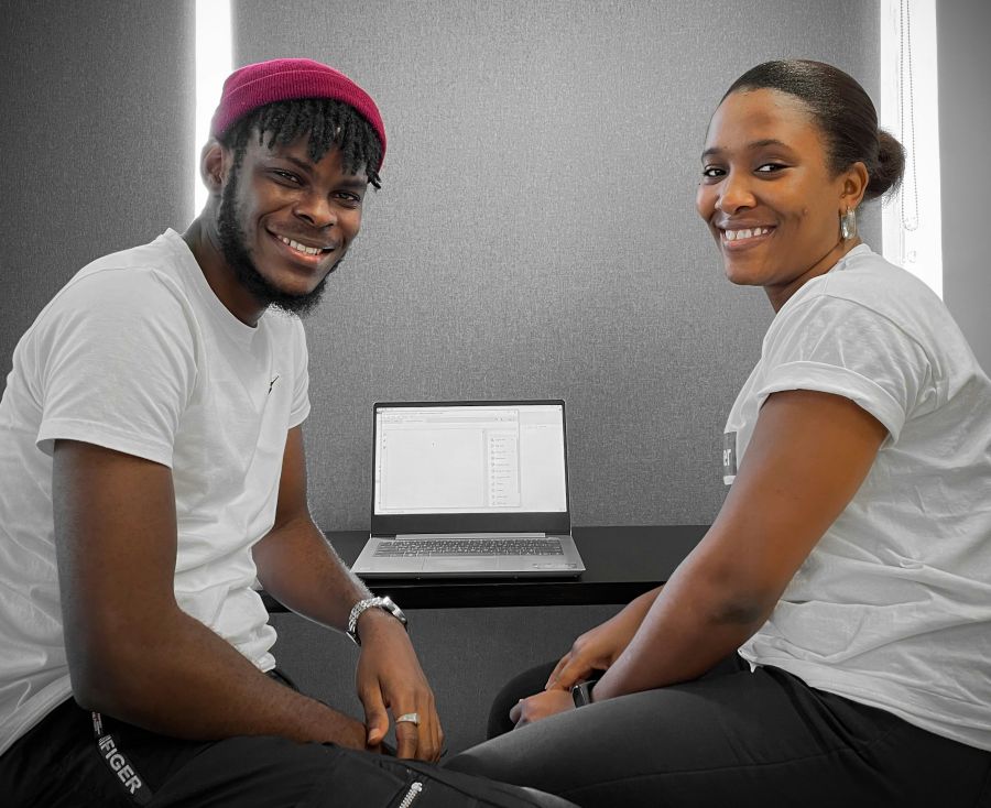 Two young people with medium skin tone are sitting in front of a laptop and are turned sideways to smile at the camera. | Deux jeunes gens au teint moyen sont assis devant un ordinateur portable et se tournent de profil pour sourire à la caméra.
