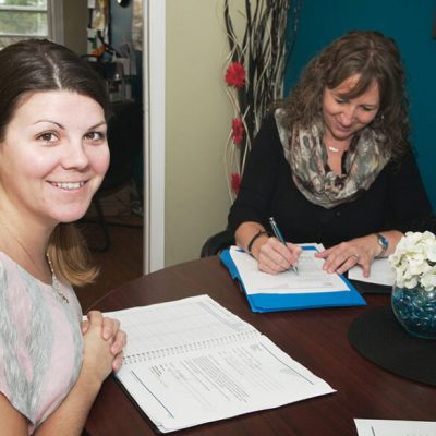 CCA-Promo-2016-001-resize-585×585 Two people sitting at a table in an office with papers on the table. One person is looking at the camera and smiling while the other is writing.