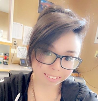 Creative Employment Options Participant, Rebecca A young woman with short dark brown hair with long bangs pushed to the side, brown eyes, and large dark stylish glasses who is smiling and wearing a black sweater with a hood.