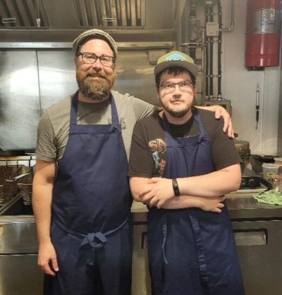 Two men standing in a commercial kitchen wearing long blue aprons. One is a bit taller, has a beard and is wearing a beret and a green t-shirt, and has his left arm around the shoulders of the other man. The second man is wearing glasses, a baseball cap, and a brown t-shirt and has his arms crossed in front of him.