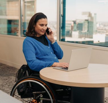 Woman with medium skin tone and shoulder length dark hair is wearing a blue sweater. She is sitting in a wheelchair at a round table in an office setting, looking at a laptop and speaking on a cellphone.