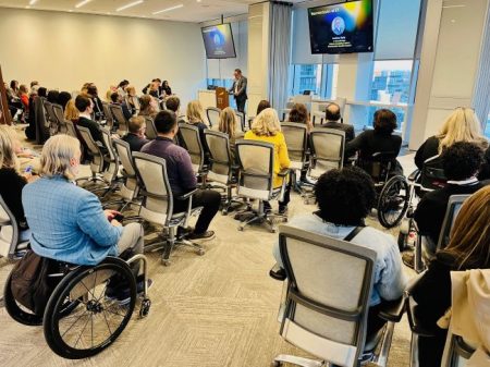 Diverse people are sitting on chairs and in wheelchairs in a large room at EY, watching a person speak at the front of the room, with television monitors above the person displaying information.