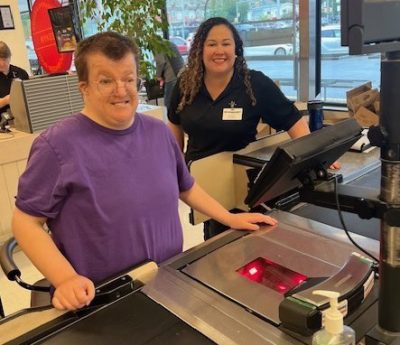 A mentee stands at a checkout counter while a mentor looks on.