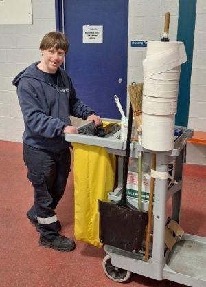 Matthew3 A man with Down syndrome has brown hair and wears a sweatshirt. He pushes a maintenance cart in an arena.