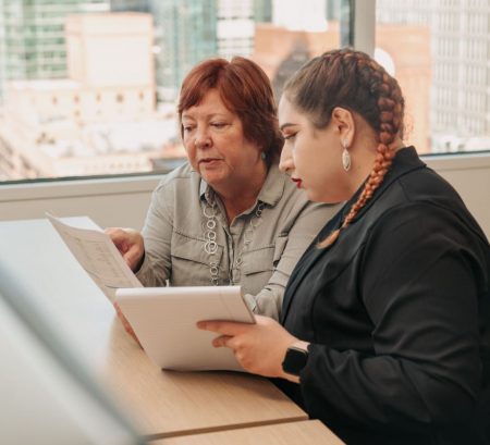 Two colleagues work together in an office. The older mentor gestures to their paper as the younger mentee follows on their notepad.