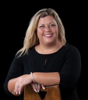 Photo - Tammy Morris. A woman with light skin tone and long blond hair wearing a black scooped-neck top is smiling and is leaning with her hands on a piece of wood.