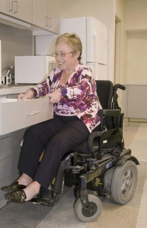 cdc-FSRxDhA5x5s-unsplash Woman with light skin in business dress in a wheelchair reaching into a drawer in a workplace kitchen.