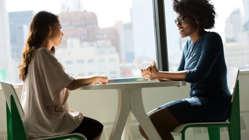 Two women, one with long hair and medium skin and one with short curly hair and dark skin, are sitting at a table beside a window at work and talking.