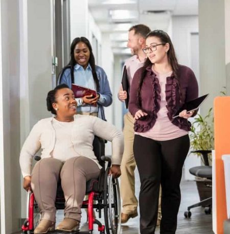 Four people are walking together in a workplace, including two women who are Black, one of whom is in a wheelchair, and one woman and one man.