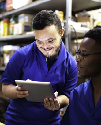 Photo: A person with Down syndrome who is standing is smiling and showing a tablet to a person beside him with dark skin who is sitting.