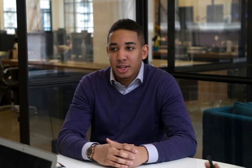 jopwell-collection-unsplash Person with medium skin and short dark hair wearing blue shirt and sweater speaks while sitting at a table in an office.