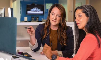 two people office unsplash In an office, a service provider with long hair and light skin points to something on a laptop to a client with long hair and medium skin.